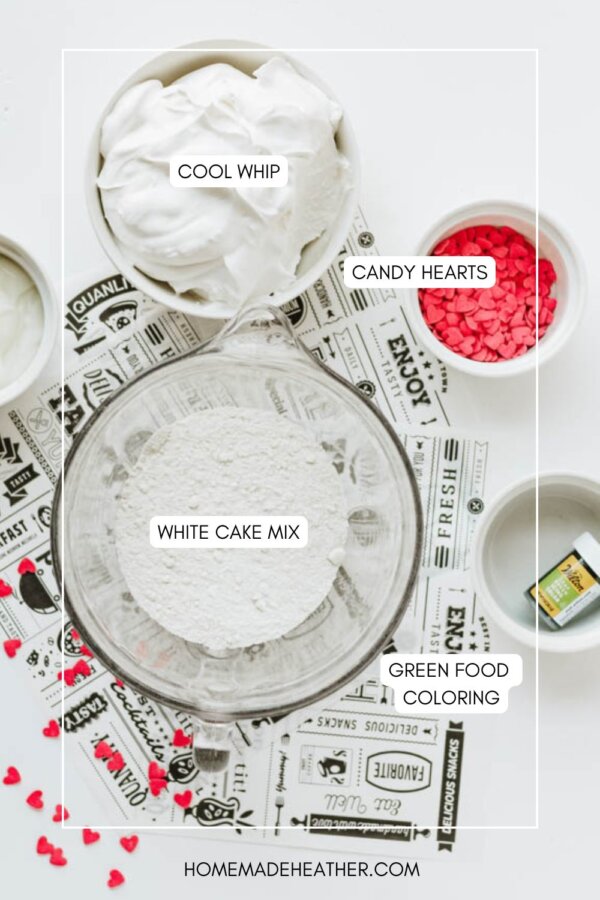 A flat lay of ingredients for a Grinch cookie dip recipe on a white surface, including a bowl of Cool Whip, white cake mix, small bowls of candy hearts, green food coloring, and a printed placemat underneath.