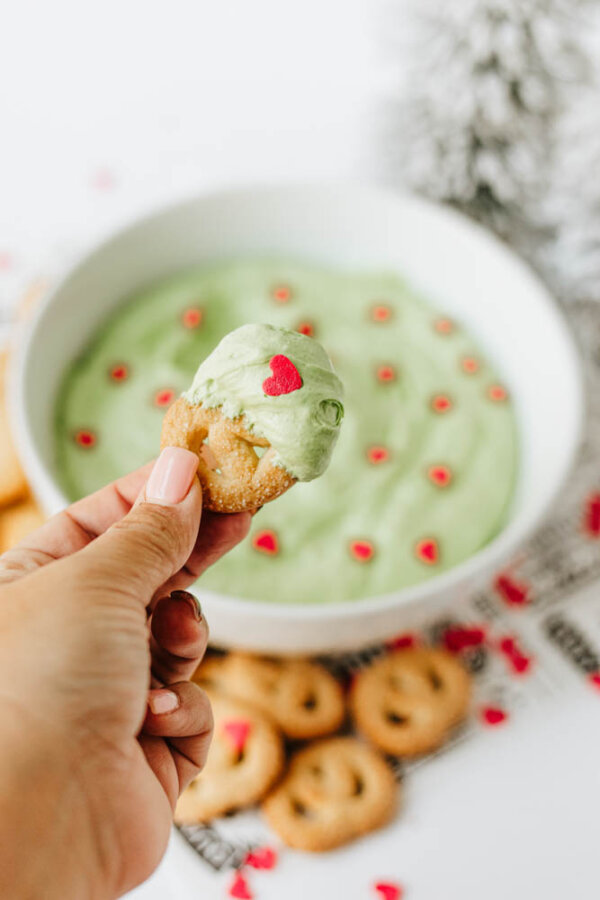 A hand holds a cookie dipped in light green cream with a red heart sprinkle. In the background, a bowl of green dip topped with small red heart sprinkles and more cookies are visible.