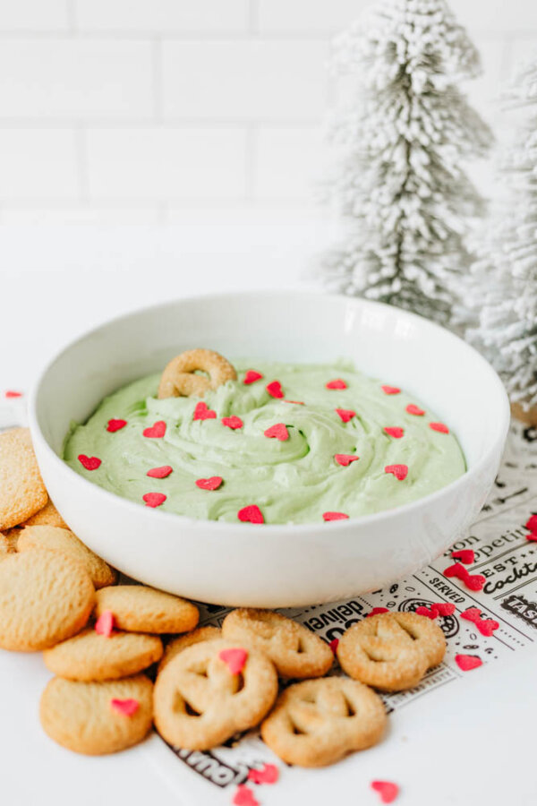 A white bowl filled with green dip topped with red heart-shaped sprinkles is surrounded by cookies and pretzels. Frosted decorative trees are in the background.