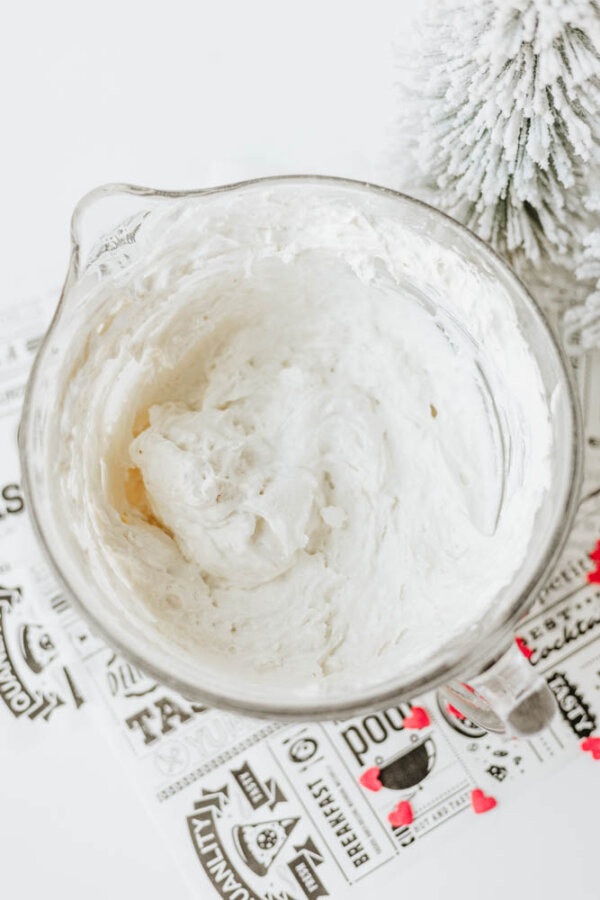 A glass mixing bowl filled with fluffy white whipped cream or frosting sits on a patterned surface next to a small, snowy artificial pine tree.