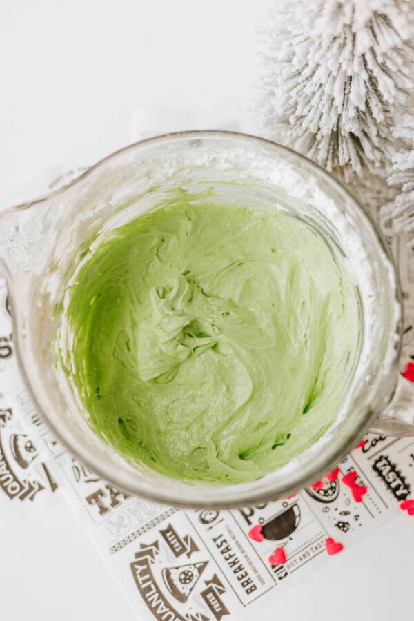 A glass mixing bowl filled with light green frosting sits on a patterned paper next to a white, snowy artificial Christmas tree.