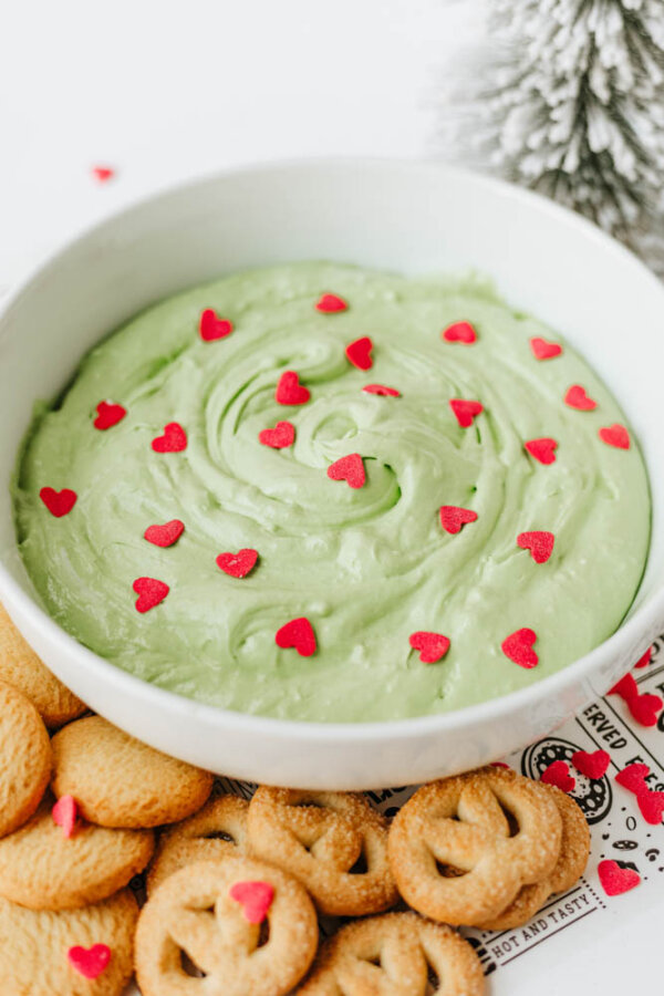 A bowl of light green dip topped with small red heart sprinkles, surrounded by round cookies and pretzel-shaped cookies on a white surface.
