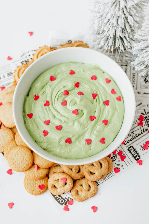 A white bowl filled with green dip topped with small red heart sprinkles, surrounded by round and pretzel-shaped cookies on a festive paper, with a snowy decorative tree in the background.