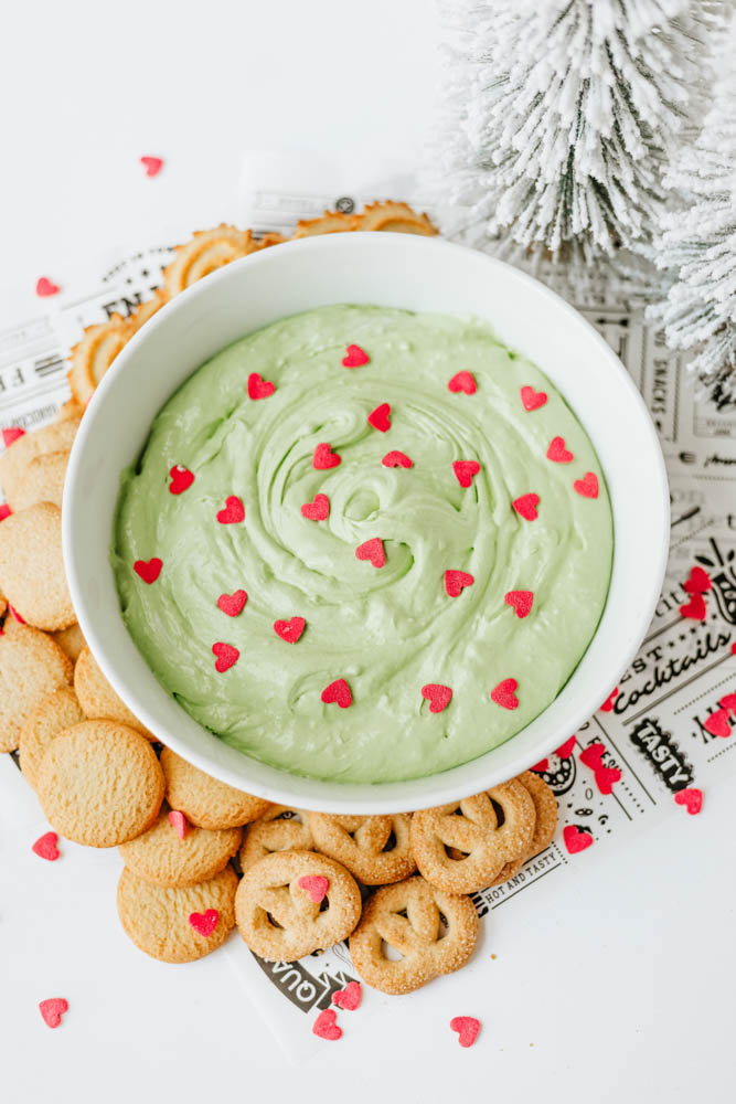A white bowl filled with green dip topped with small red heart sprinkles, surrounded by round and pretzel-shaped cookies on a festive paper, with a snowy decorative tree in the background.