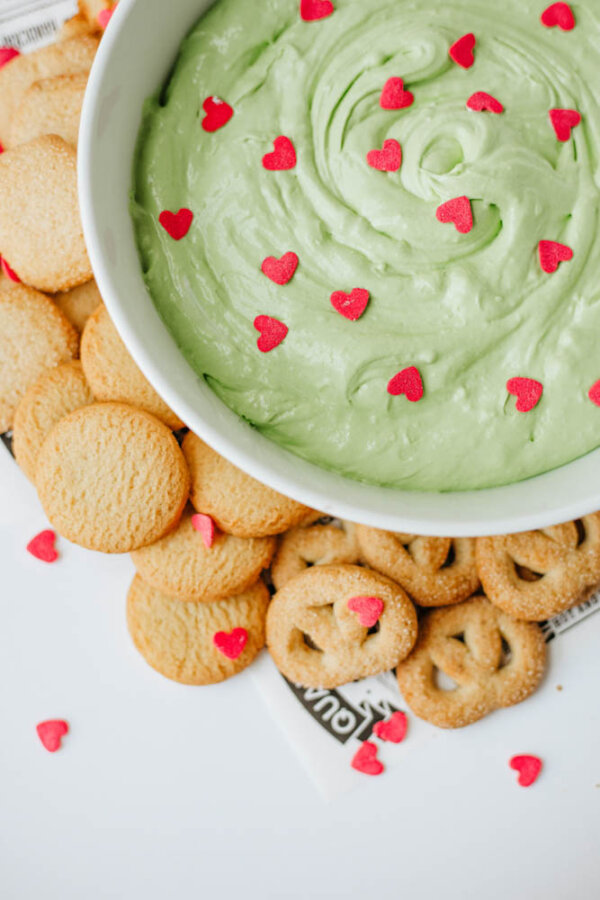 A bowl of green dip topped with small red heart sprinkles, surrounded by round cookies and pretzels on a white surface. Some heart sprinkles are scattered on the snacks and table.
