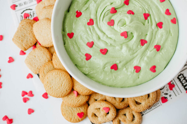 A white bowl filled with green dip topped with small red heart-shaped sprinkles, surrounded by various cookies on a white surface.