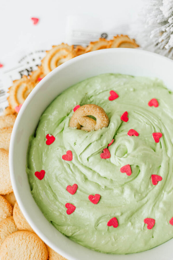 A white bowl filled with light green dip, topped with small red heart sprinkles and a round cookie in the center, surrounded by more cookies on the side.
