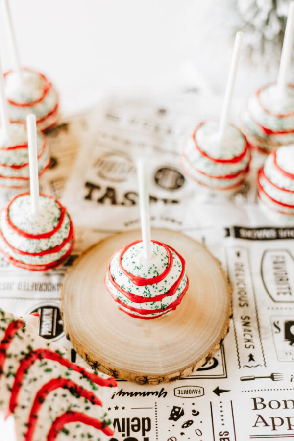 Round cake pops decorated with white frosting, red and green sprinkles, and red icing stripes are displayed on sticks. One is placed on a wooden slab, with others arranged on printed paper in the background.