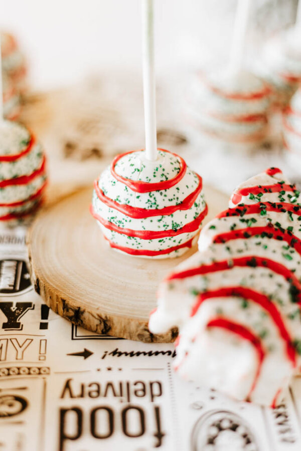 A cake pop decorated with white icing, red drizzle, and green sprinkles sits on a wooden slice, with similarly decorated treats in the background on a patterned surface.