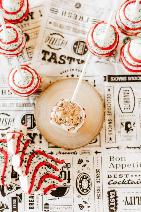 Festive cake pops with red, green, and white sprinkles are arranged on a wooden slice and decorative paper with food-themed words. One cake pop is displayed upright in the center.