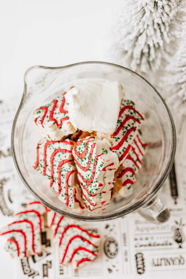 A glass mixing bowl filled with Christmas-themed cookies decorated with red and green icing and sprinkles, topped with a dollop of white cream, placed on a festive background near a snow-dusted tree.