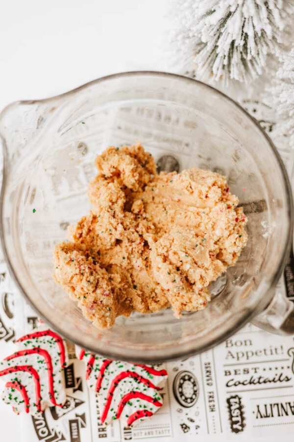 A glass mixing bowl filled with raw cookie dough sits on a decorative paper with text. Nearby are striped holiday cookies and part of a snow-dusted artificial pine tree, creating a festive baking scene.