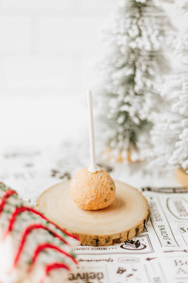 A cake pop with a light brown crumb coating sits on a wooden slice, surrounded by festive holiday decor and faux snow-covered trees in the background.