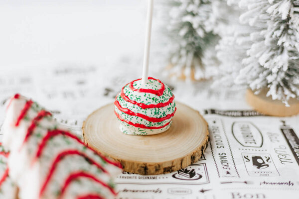 A cake pop decorated with white icing, green sprinkles, and red drizzle sits on a wooden slice. Snow-covered artificial trees are in the background, creating a festive holiday scene.