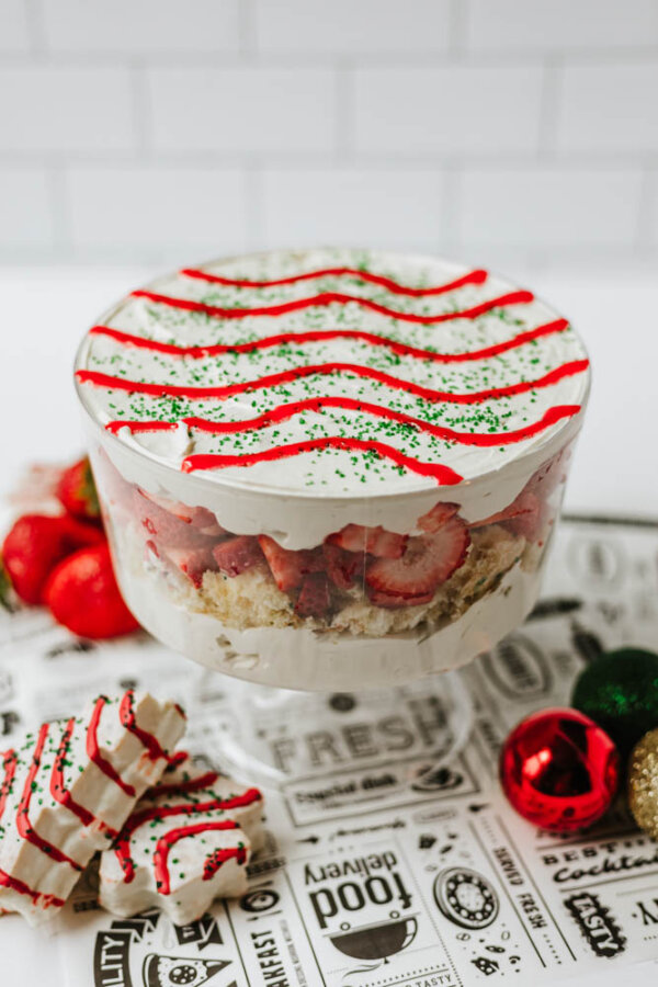 A layered trifle dessert with whipped cream, strawberries, and cake in a glass bowl, topped with red icing stripes and green sprinkles. Surrounded by peppermint treats and festive Christmas ornaments.