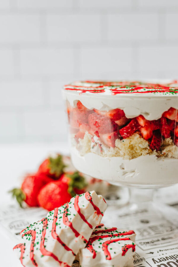 A trifle dessert in a glass bowl with layers of whipped cream, sponge cake, and fresh strawberries. In front, there are decorated white chocolate-covered treats and whole strawberries on a printed surface.