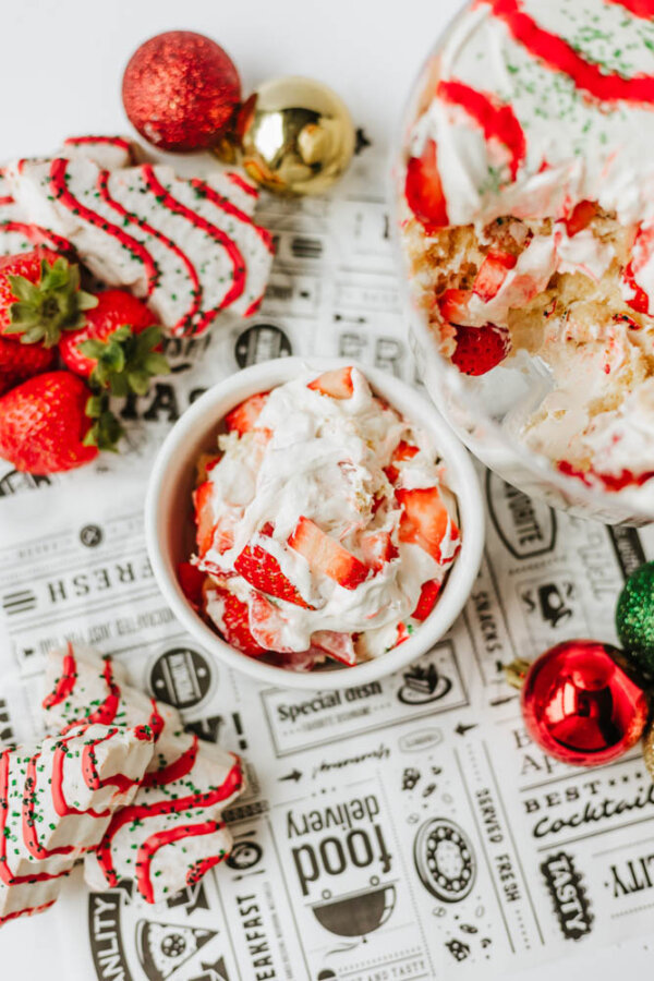 A bowl of strawberries topped with whipped cream sits beside a larger dessert with similar toppings, festive cookies, fresh strawberries, and colorful Christmas ornaments on a decorative paper.
