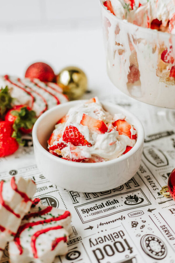 A white bowl filled with a creamy dessert topped with fresh strawberries sits on a printed tablecloth, with more strawberries and what appears to be a trifle dessert visible in the background.