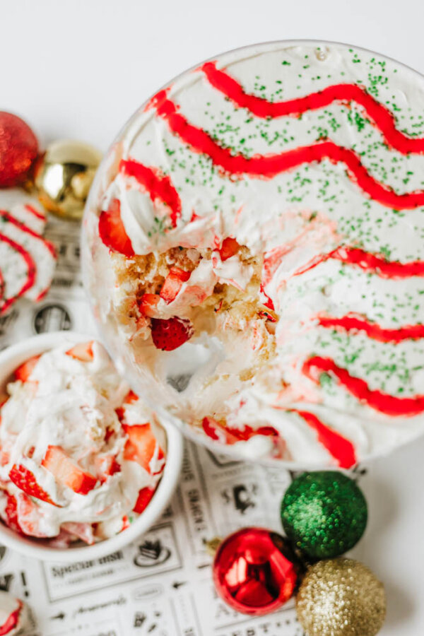 A festive trifle dessert with layers of whipped cream and strawberries, decorated with red syrup and green sprinkles, sits beside Christmas ornaments and a small dish of the dessert.