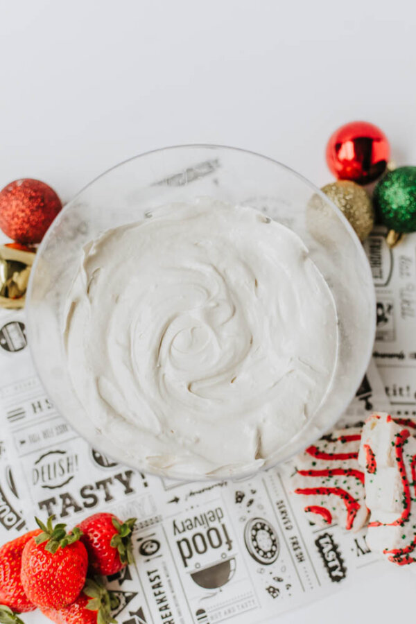 A glass bowl filled with creamy white dip, placed on a patterned paper with strawberries, a decorated cookie, and festive ornaments nearby.