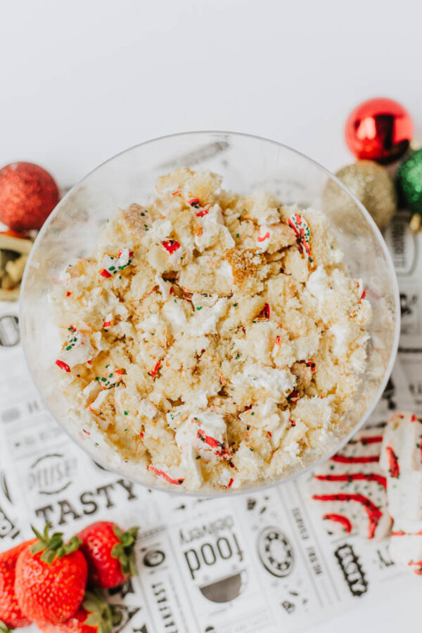 A glass bowl filled with crumbled cake and colorful sprinkles sits on a printed placemat, with strawberries and festive ornaments nearby.