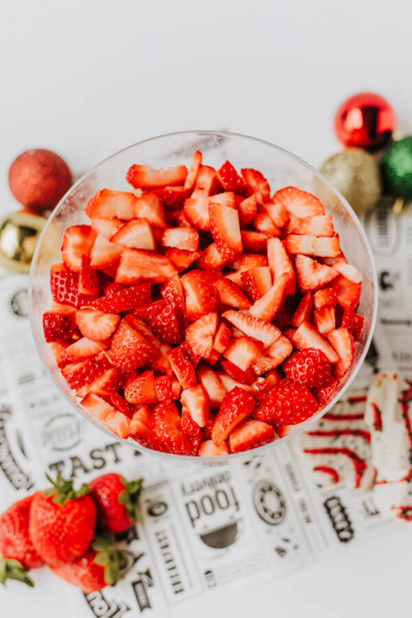 A glass bowl filled with sliced strawberries sits on a decorative napkin, with whole strawberries and colorful holiday ornaments nearby.