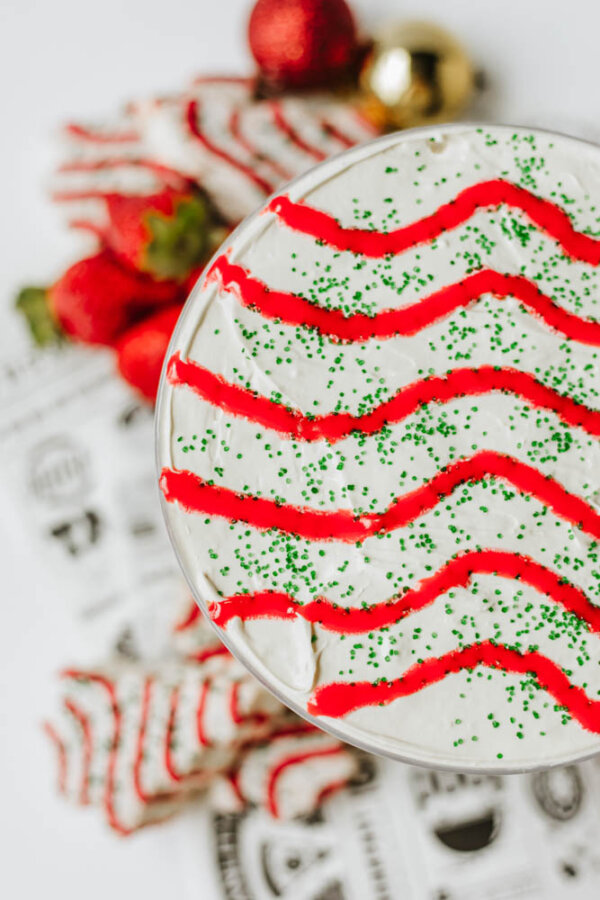 A close-up of a holiday dessert with white frosting, red zigzag icing, and green sprinkles. Strawberries and decorated treats are blurred in the background, giving a festive appearance.
