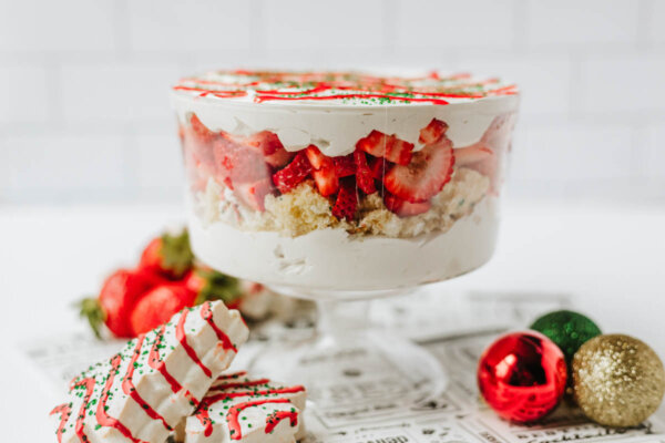 A layered trifle dessert with whipped cream, cake pieces, and fresh strawberries in a glass bowl sits on a newspaper print, surrounded by strawberries, decorated cookies, and shiny red and green ornaments.