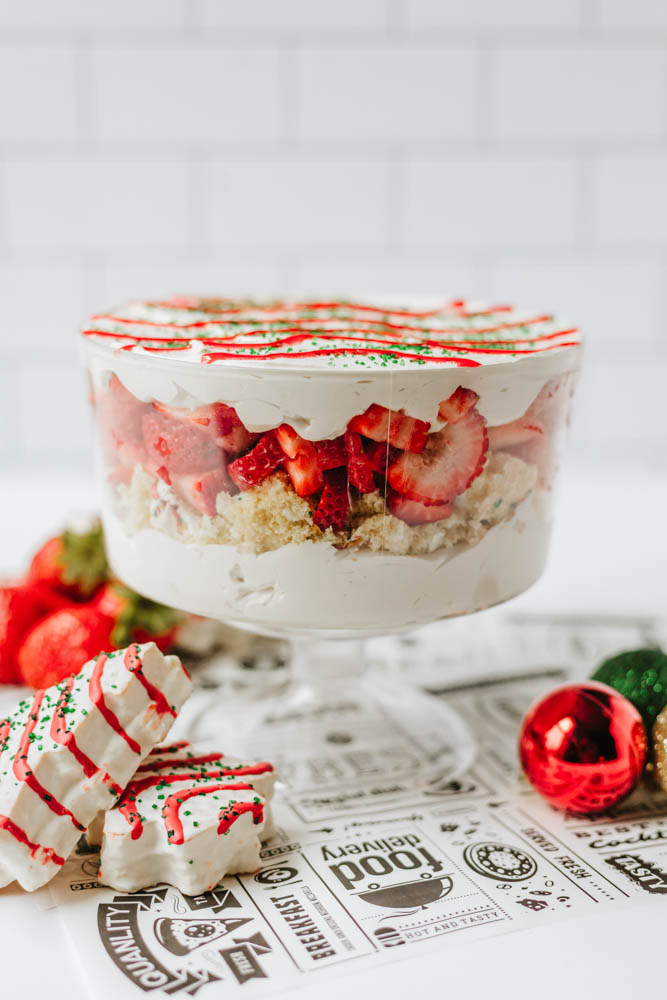 A layered trifle dessert with whipped cream, cake pieces, and sliced strawberries in a glass bowl, topped with red drizzle and green sprinkles. Decorated cookies and Christmas ornaments are beside the bowl.