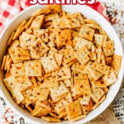 A bowl filled with seasoned saltine crackers, topped with red pepper flakes, sits on a patterned surface with a red and white checkered cloth nearby. Text reads โfirecracker saltinesโ and โSO GOODโ with a website link at the bottom.