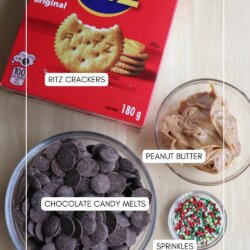 A top-down view of ingredients for a snack: a box of Ritz crackers, a bowl of chocolate candy melts, a bowl of peanut butter, and a small bowl of colorful sprinkles on a wooden surface.