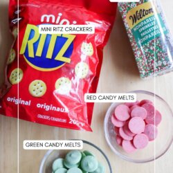 A bag of Mini Ritz crackers, a container of Wilton sprinkles, a bowl of red candy melts, and a bowl of green candy melts are arranged on a light-colored surface.