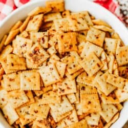 A bowl filled with seasoned square saltine crackers is shown on a table with a red checkered cloth. Text at the top reads โFirecracker Saltinesโ and at the bottom โhomemadeheather.comโ.