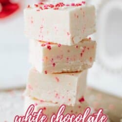 Three pieces of white chocolate fudge with red and pink sprinkles are stacked on a wooden surface. Text reads Valentines Day white chocolate fudge recipe homemadetheather.com.