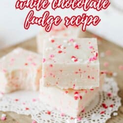 Three pieces of white chocolate fudge with red and pink sprinkles are stacked on a wooden board with a lace doily. Text reads “Valentine’s Day white chocolate fudge recipe – so good!”.