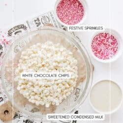 A glass bowl filled with white chocolate chips, a small bowl of sweetened condensed milk, and two bowls of red and pink festive sprinkles are arranged on a white surface.