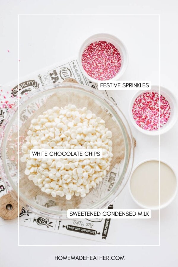 A glass bowl filled with white chocolate chips, a small bowl of sweetened condensed milk, and two bowls of red and pink festive sprinkles are arranged on a white surface.