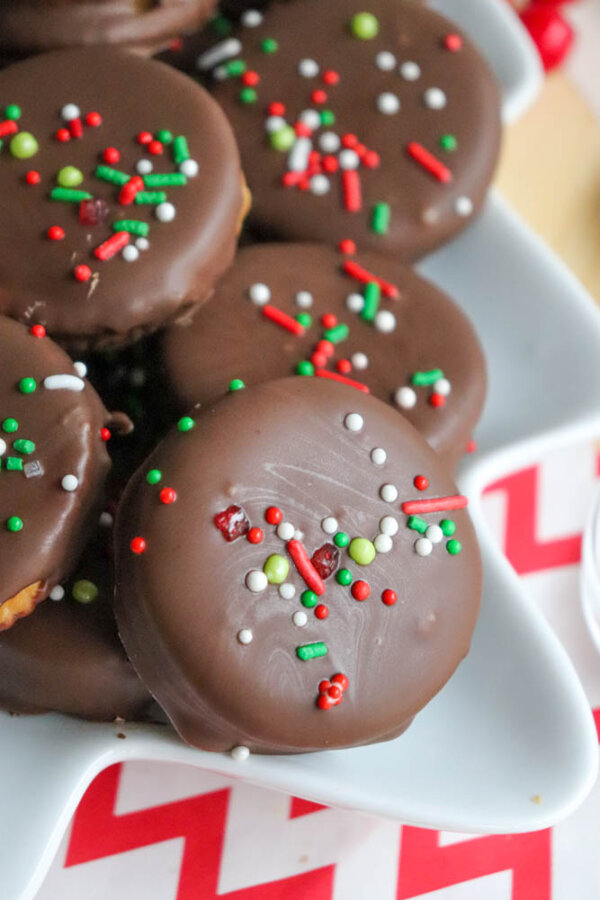 A plate of chocolate-covered cookies topped with red, green, and white sprinkles, arranged on a white dish with a red and white chevron-patterned tablecloth underneath.