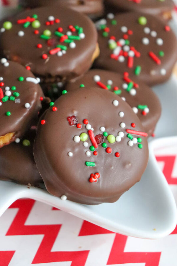 Chocolate-covered cookies topped with red, green, and white sprinkles are arranged on a white plate with a red and white chevron-patterned surface underneath.