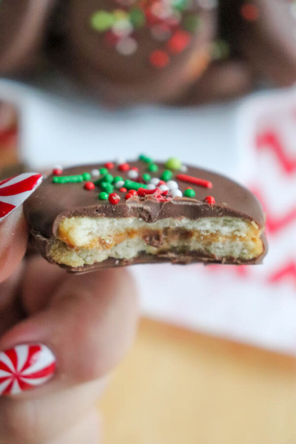 A hand with festive nail art holds a chocolate-covered cookie with a bite taken out, showing layers of caramel and cracker, topped with green, red, and white sprinkles.