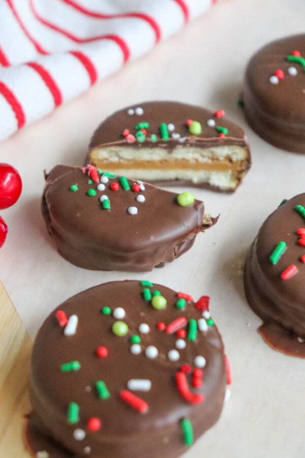 Four chocolate-covered sandwich cookies with colorful red, white, and green sprinkles. One cookie is cut in half, showing layers of cream and cookie inside. A red-and-white striped cloth is in the background.