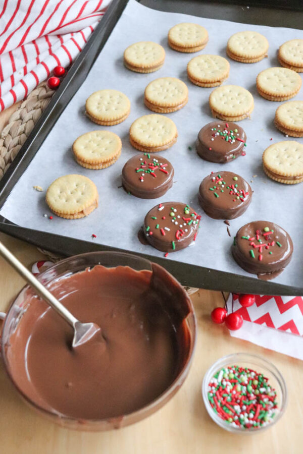 A baking tray with Ritz crackers, some dipped in chocolate and topped with holiday sprinkles, sits beside a bowl of melted chocolate and a small container of sprinkles on a wooden table.