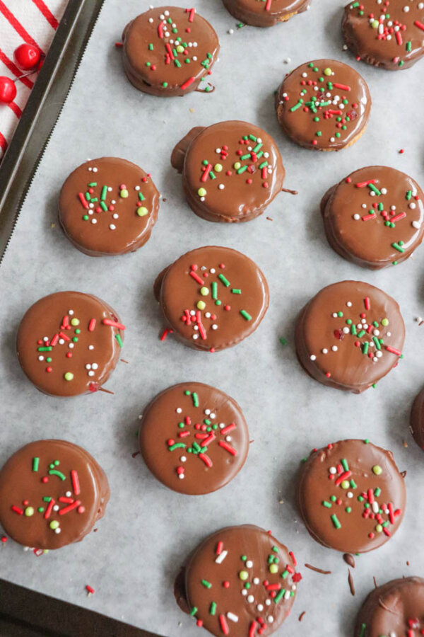 Round chocolate-covered cookies topped with red, green, and white sprinkles are arranged neatly on a parchment-lined baking sheet. Some chocolate coating and sprinkles are scattered around the cookies.