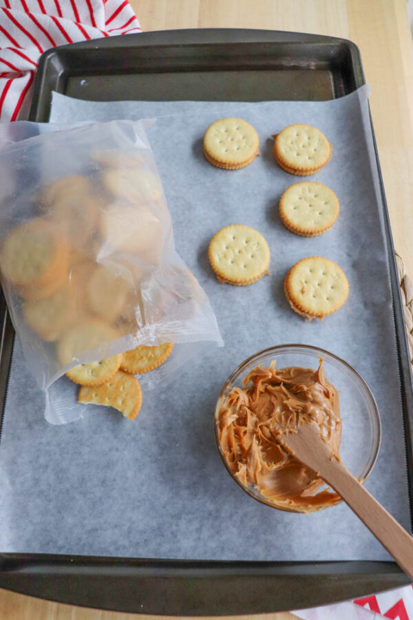 A baking tray lined with parchment paper holds several round crackers, an open plastic bag with more crackers, and a glass bowl of peanut butter with a wooden spatula.