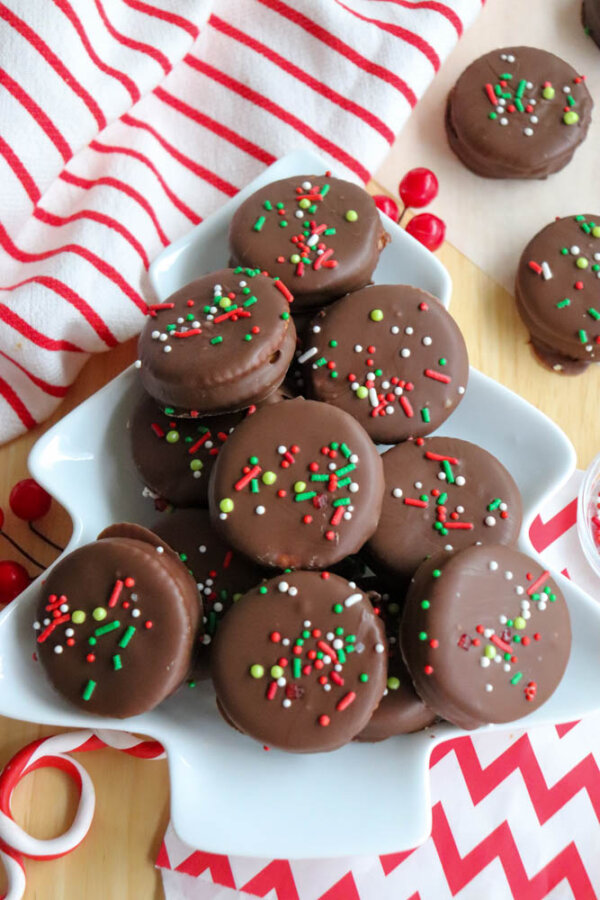 A white, tree-shaped plate holds chocolate-covered sandwich cookies topped with red, green, and white sprinkles. Red and white striped napkins decorate the festive setting.