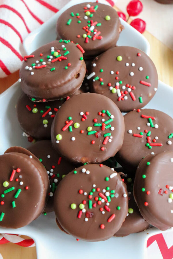 A plate of round chocolate-covered cookies topped with festive red, green, and white sprinkles, arranged on a white dish with a striped cloth and red candies nearby.