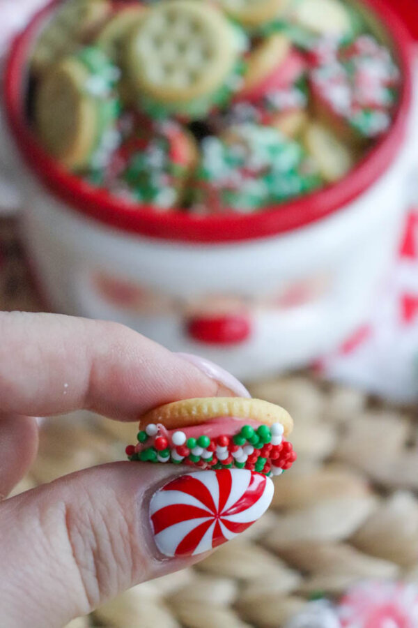 A hand with a red and white peppermint swirl nail design holds a mini sandwich cookie coated with red, white, and green sprinkles. A festive bowl filled with similar cookies is blurred in the background.