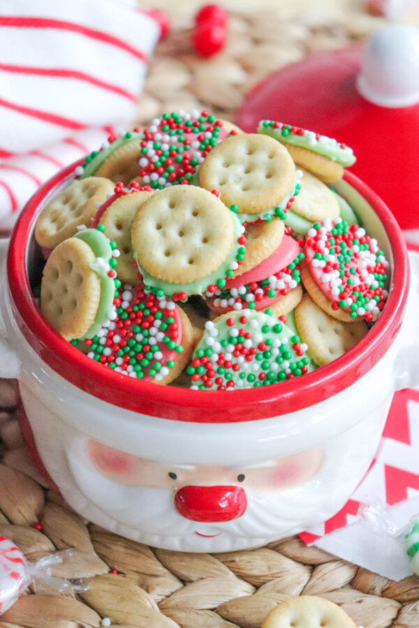 A festive jar shaped like Santaโs face is filled with round cracker sandwiches dipped in white and green chocolate and coated with red, green, and white sprinkles. A red and white towel and dish are in the background.