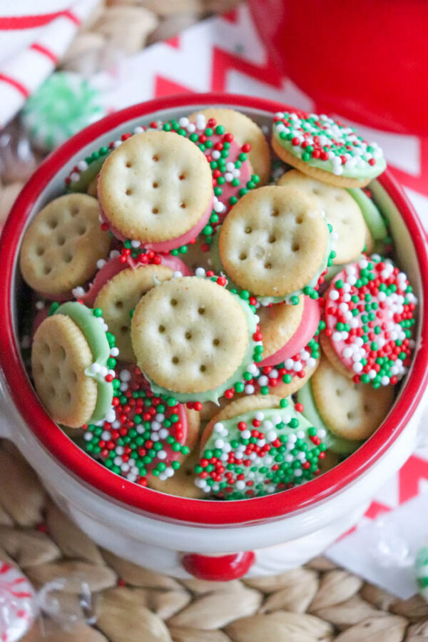 A bowl filled with mini sandwich cookies coated in red, green, and white sprinkles, decorated for the holidays, sits on a woven placemat and red-and-white patterned surface.