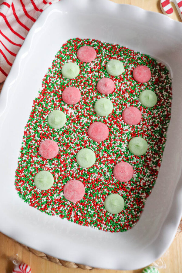 A white dish filled with red, green, and white sprinkles, topped with alternating pink and green candy melts arranged in rows. A red and white striped cloth is partially visible in the corner.